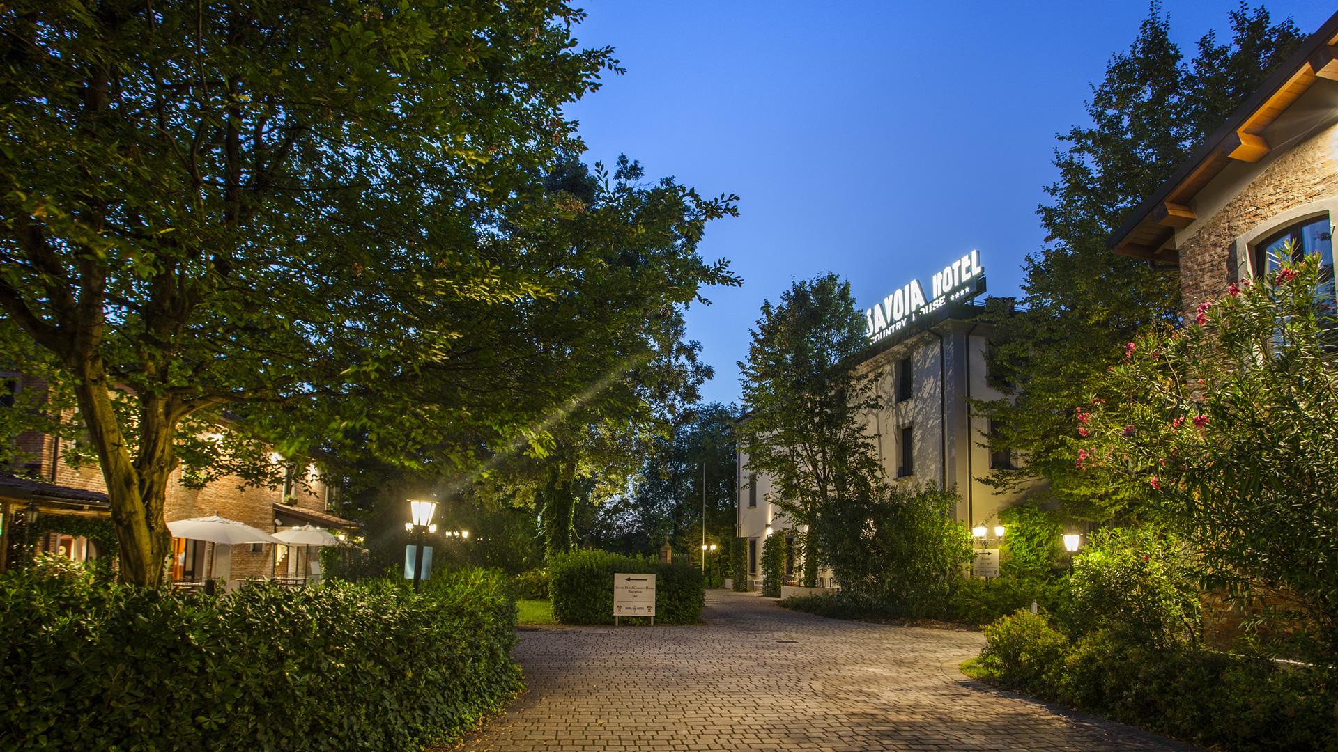 Savoia Hotel Country House con giardino verde e panchina bianca di sera.