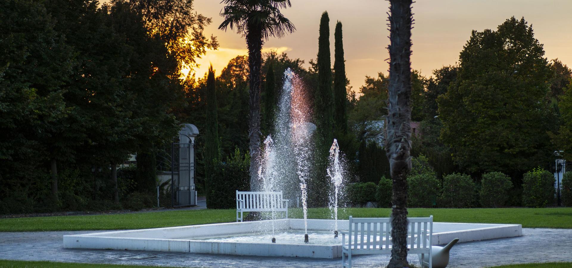 Fontana con panchine bianche in un parco al tramonto.