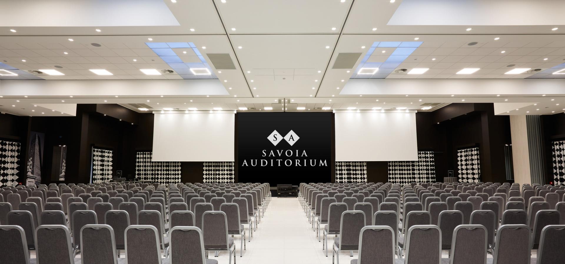Un moderno auditorium con sedie grigie e schermi per presentazioni.