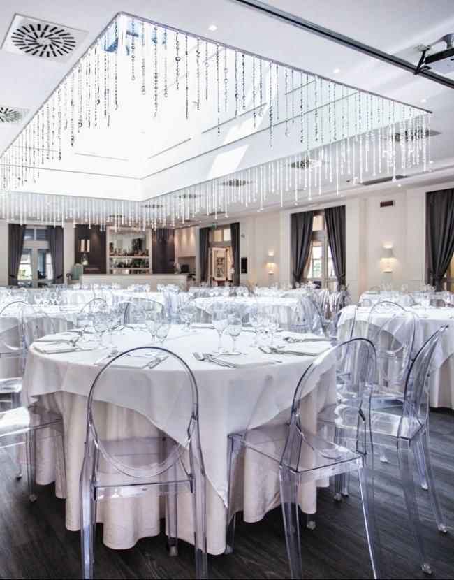 Elegant hall with set round tables and transparent chairs, illuminated by modern chandeliers.