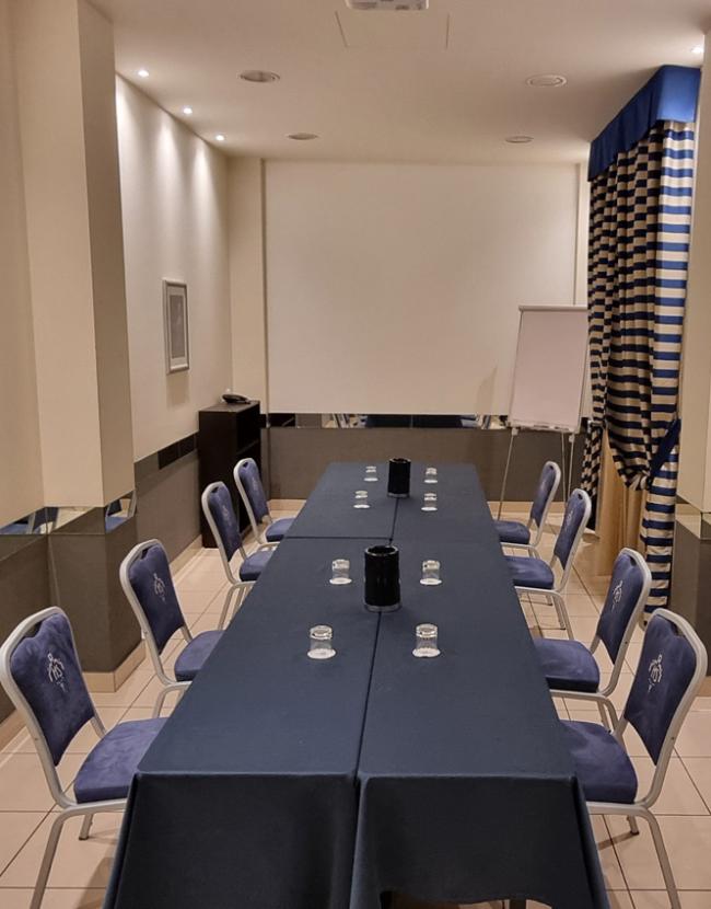 Sala riunioni con tavolo rettangolare, sedie blu e schermo per proiezioni.