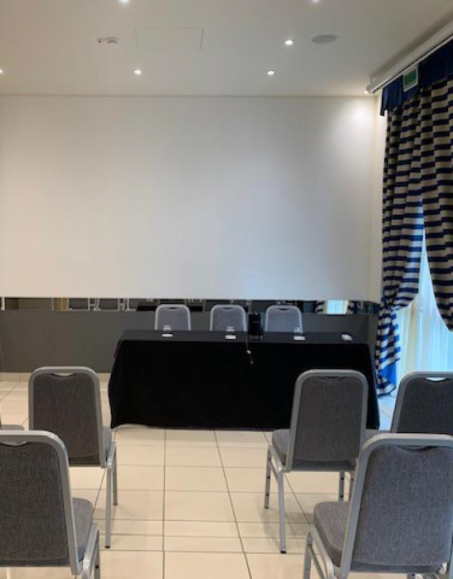 Sala conferenze con sedie, tavolo, schermo e tende a righe.