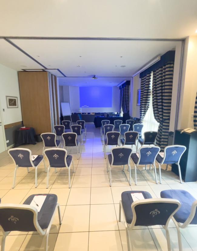 Sala conferenze con sedie blu, schermo di proiezione e lavagna bianca.