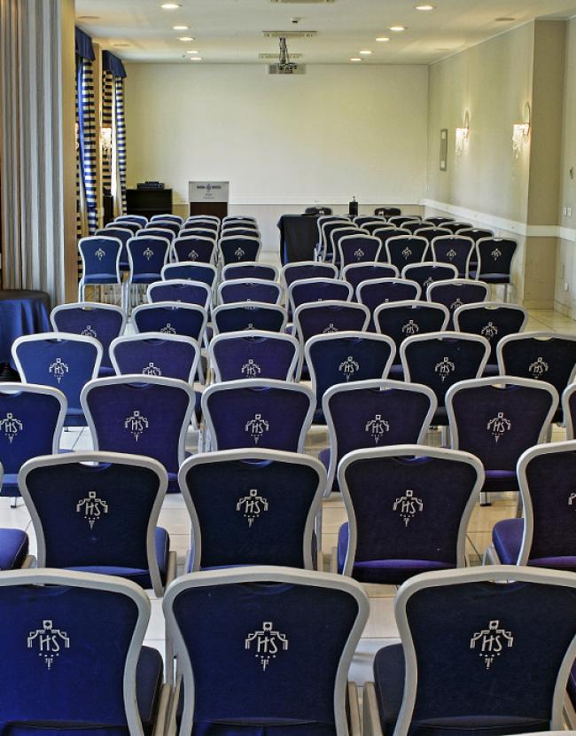 Sala conferenze con sedie blu e bianche, tende a righe, e proiettore.