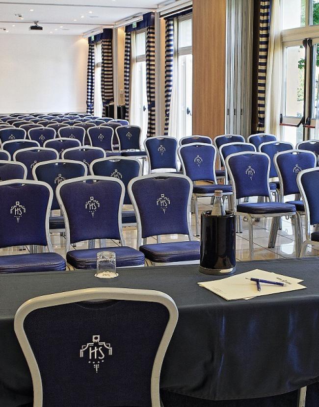 Sala conferenze con sedie blu, tavolo con microfono, bottiglia e fogli.
