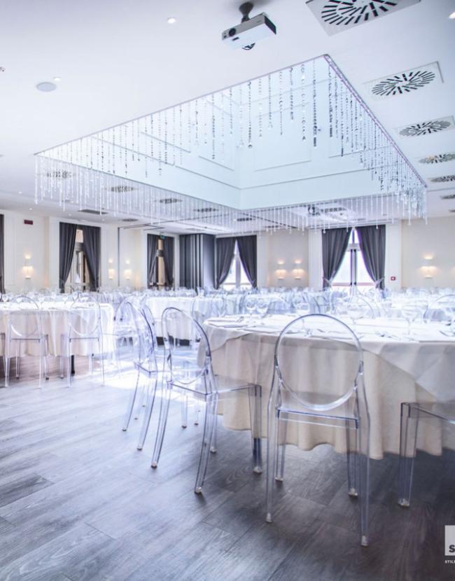 Sala elegante con tavoli rotondi, sedie trasparenti e decorazioni a soffitto.