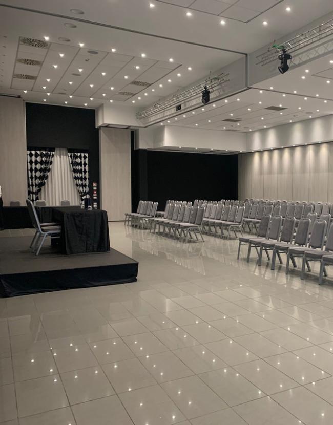 Sala conferenze moderna con sedie grigie, palco e illuminazione a soffitto.