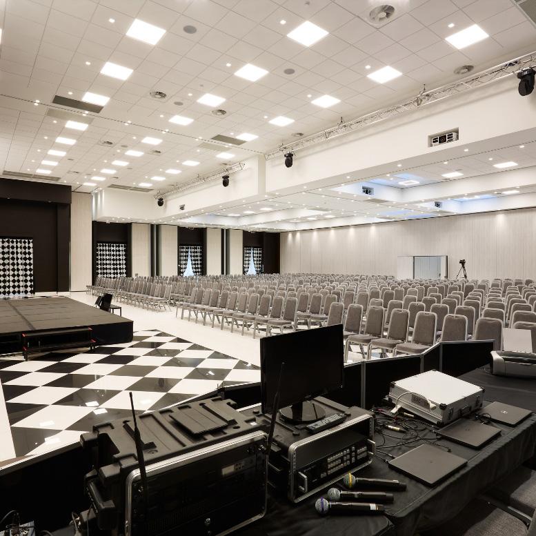 Sala conferenze con palcoscenico, sedie e attrezzatura audiovisiva.